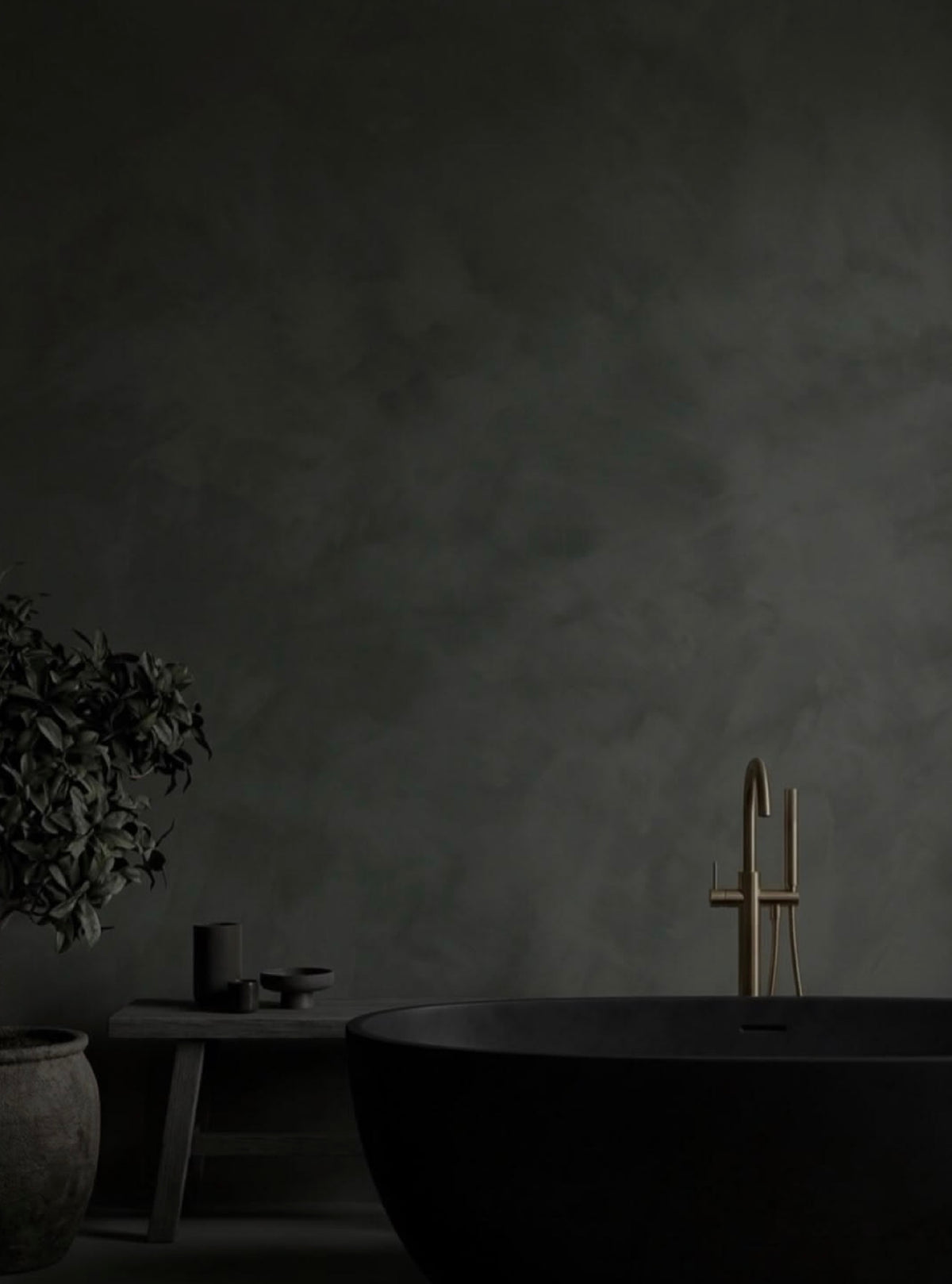 A dark freestanding bathtub in a minimalist bathroom setting with a matte dark gray plaster wall, a small wooden bench with candles, and a potted plant on the left. A tall matte brass faucet is mounted on the wall above the tub.